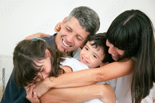 Young Caucasian family hugging and smiling. Beautiful brunette mother embracing cute son, lovely daughter and happy husband with love. Parenthood, fun, children and togetherness concept