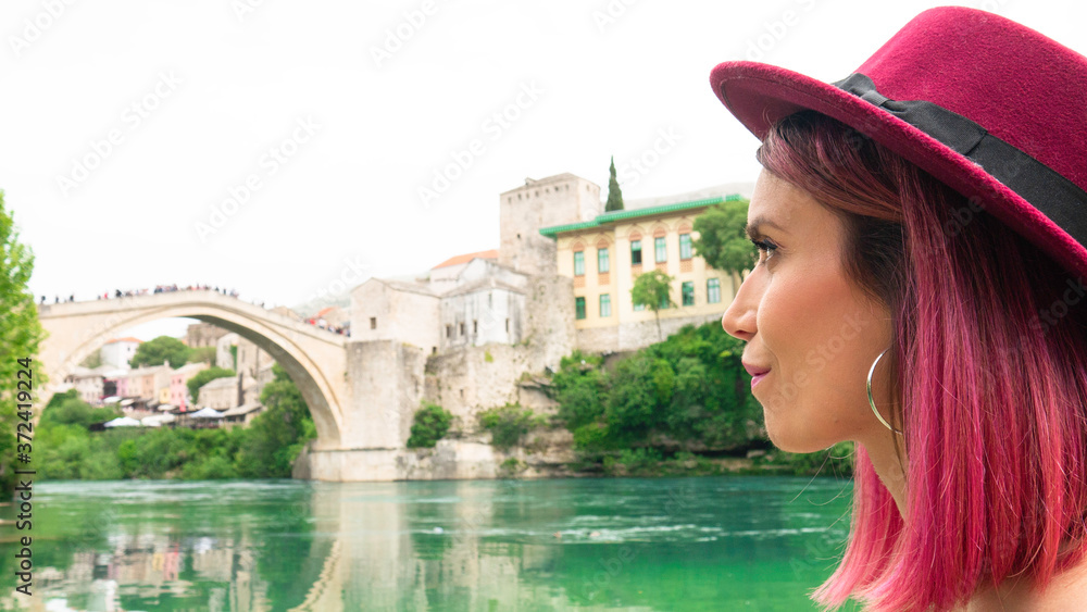Portrait of a young happy woman with pink hair and a hat who observes the beauty of Mostar and the Old Bridge over the river Neretva, Bosnia and Herzegovina. Tourists sightseeing Mostar.