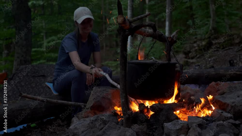 A woman watches the fire while cooking in pot on a campfire at dusk A woman watches the fire while cooking in pot on a campfire at dusk