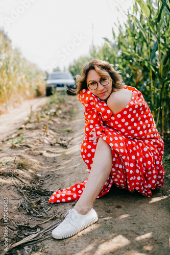 Cheerful young caucasian female with short dark wavy hair in glasses wrapped in red and white coverlet keeps calm in the cornfield in the weekends