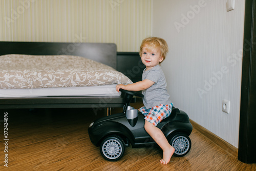 Picture of cute little caucasian boy with short fair hair in grey t-shirt and shorts rides a toy black car in parent's bedroom
