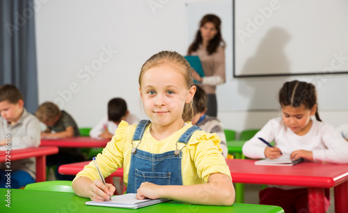 Portrait of little assiduous girl with pen and notebook at lesson in elementary school..