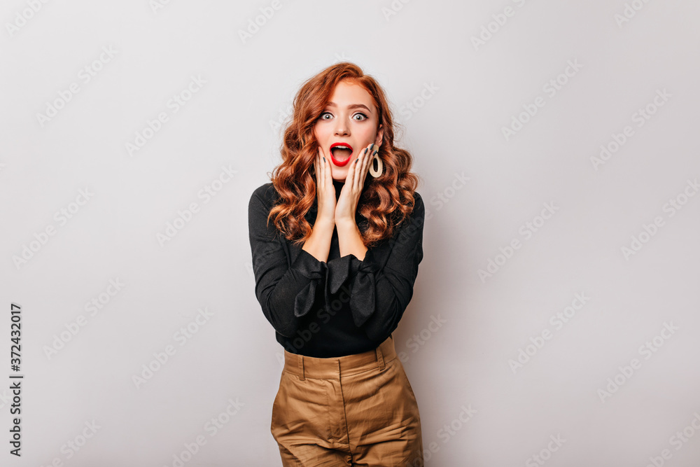 Shocked curly woman in big golden earrings standing on light background. Studio photo of emotional red-haired girl. Shocked curly woman in big golden earrings standing on light background. Studio photo of emotional red-haired girl.
