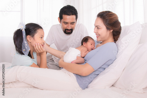 Happy family concept, father and daughter sit on bed excited looking new member in family, adorable newborn sleep on mother chest, mom hold infant embracing and caring while look daughter with love