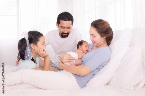 Happy family concept, father and daughter sit on bed excited looking new member in family, adorable newborn sleep on mother chest, mom hold infant embracing and caring while look daughter with love