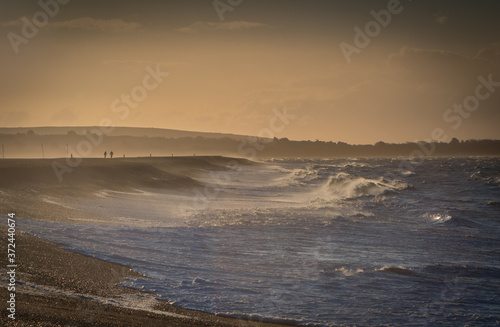 Stormy beach scene with two silhouetted people along Hurst Spit, Milford-on-Sea, Hampshire UK. Contrast of blue sea against early morning light as sun moves over the horizon photo