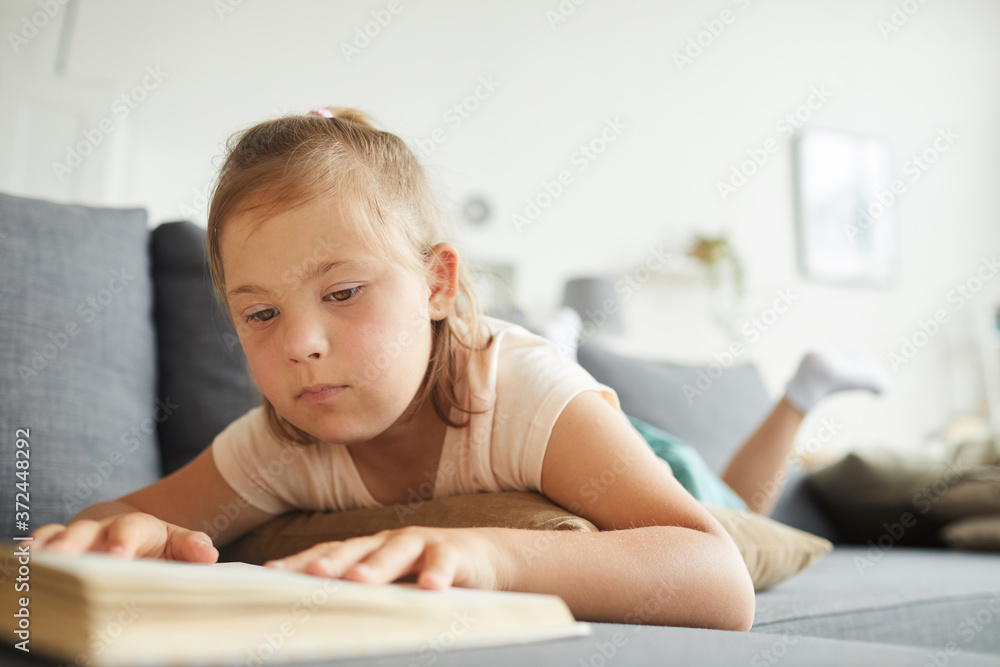 Little girl with down syndrome lying on sofa and reading a book in the living room Little girl with down syndrome lying on sofa and reading a book in the living room