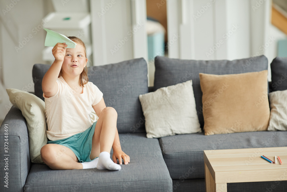 Girl with down syndrome sitting on sofa and playing with paper plane in the living room Girl with down syndrome sitting on sofa and playing with paper plane in the living room