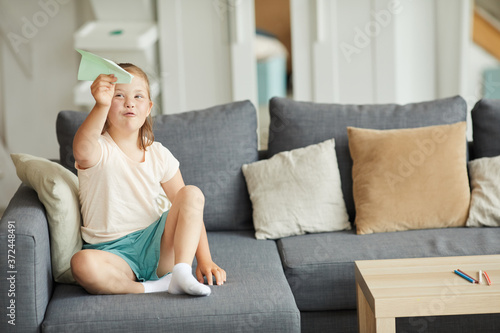 Girl with down syndrome sitting on sofa and playing with paper plane in the living room