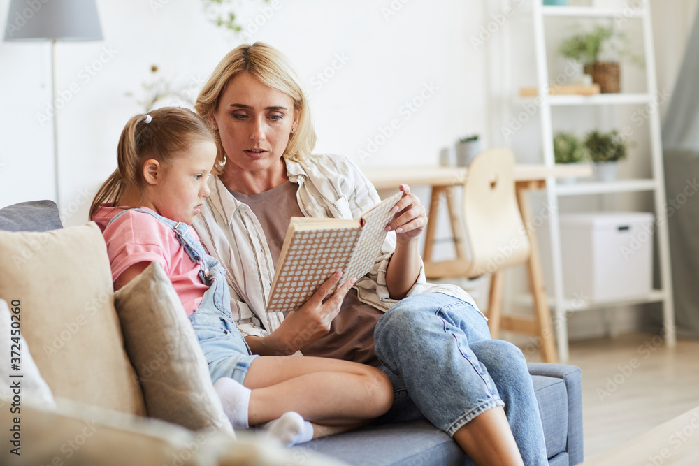 Young mother reading a book to her daughter with down syndrome while they sitting on sofa in the room