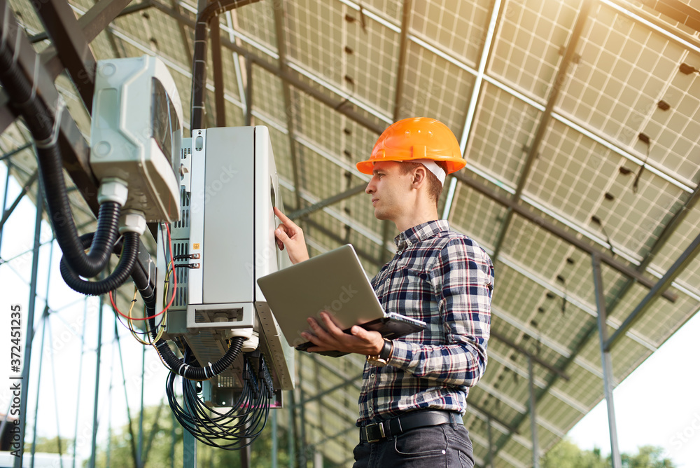 Expert is inspecting quality of solar panels. Man in hardhat standing with laptop near electrical equipment. Solar station sunny power electricity. Innovation future. Expert is inspecting quality of solar panels. Man in hardhat standing with laptop near electrical equipment. Solar station sunny power electricity. Innovation future.