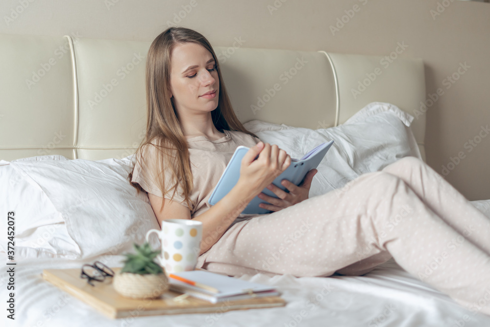 Happy and smiling relaxed cheerful woman reading notebook and working from home office on bed and drinking coffee. Blonde girl Enjoying free leisure time with book