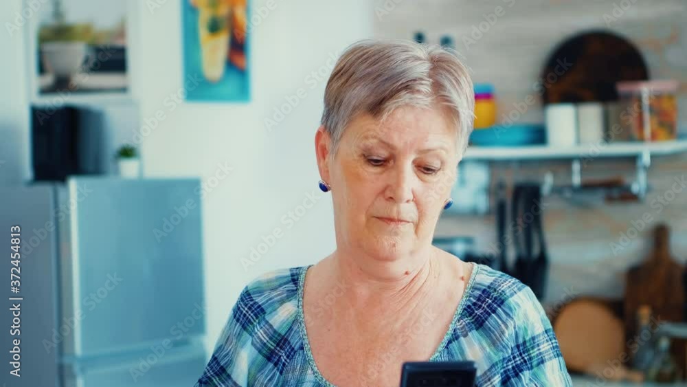 Granny looking at camera while using smartphone in kitchen during breakfast. Authentic elderly person using modern smartphone internet technology. Online communication connected to the world, senior Granny looking at camera while using smartphone in kitchen during breakfast. Authentic elderly person using modern smartphone internet technology. Online communication connected to the world, senior