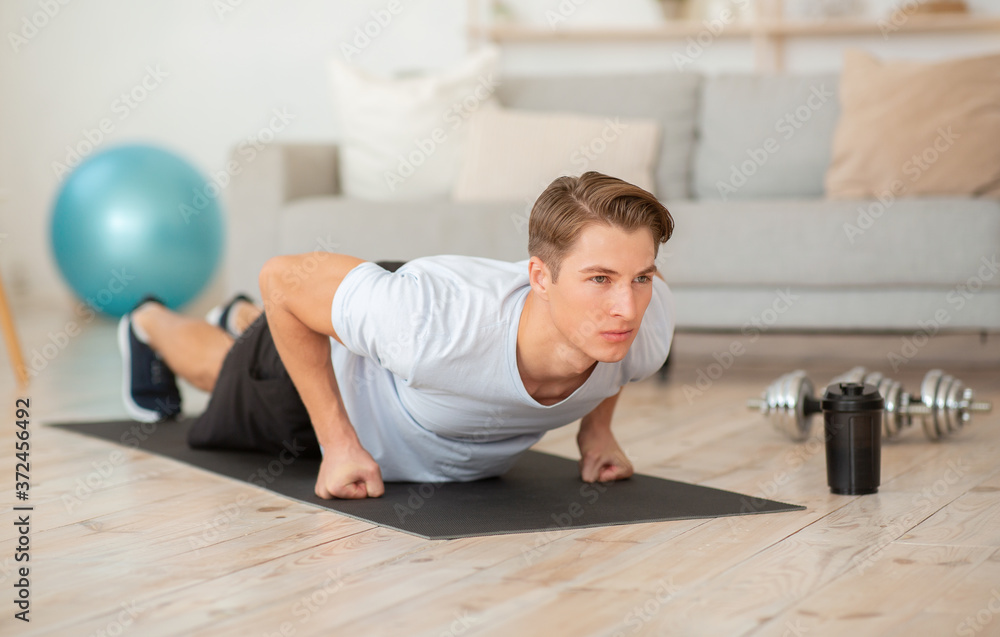 Concentrated guy push up on fists on mat in interiowith dumbbells, ball and bottle of water Concentrated guy push up on fists on mat in interiowith dumbbells, ball and bottle of water