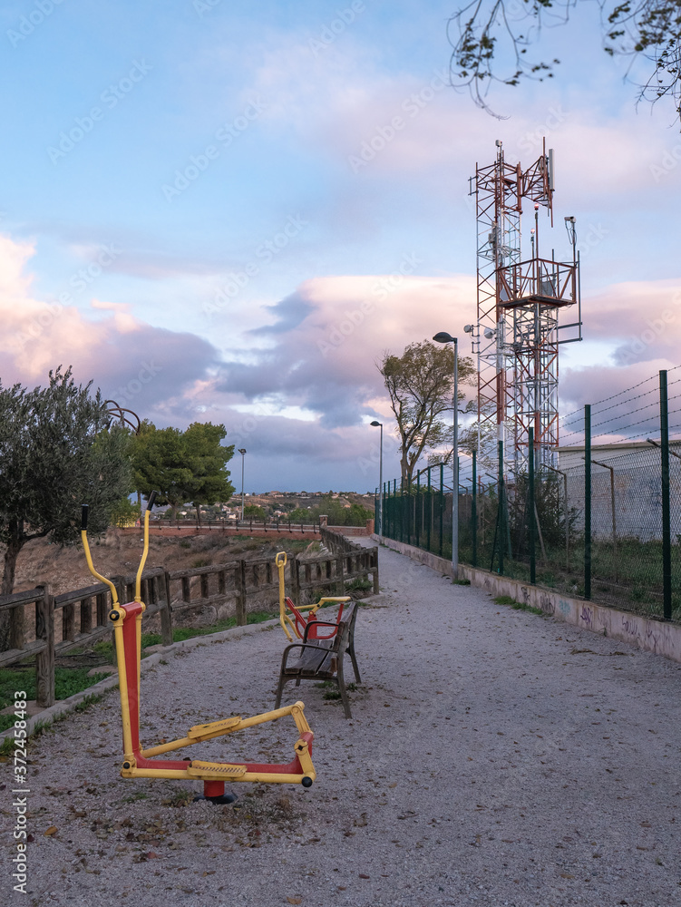 Biohealthy circuit for the elderly with two exercise equipment in a park with a wooden fence walkway and a telecommunications tower to one side during sunrise Biohealthy circuit for the elderly with two exercise equipment in a park with a wooden fence walkway and a telecommunications tower to one side during sunrise