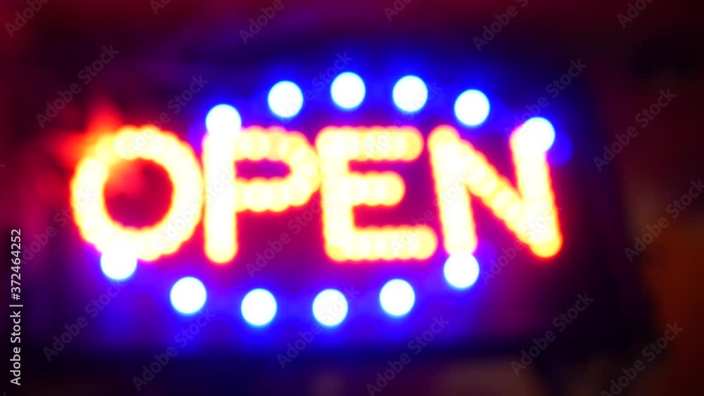 Open neon sign glowing in the dark. Vivid retro styled text at entrance on glass window. Colorful electric banner selective focus close up. Light bulbs radiance at night. Shiny illuminated lettering. Open neon sign glowing in the dark. Vivid retro styled text at entrance on glass window. Colorful electric banner selective focus close up. Light bulbs radiance at night. Shiny illuminated lettering.