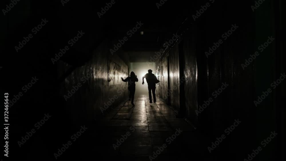 A general shot of a man and a girl running away along a straight corridor from the shadows into the light from a limping man with an ax. High quality 4k footage A general shot of a man and a girl running away along a straight corridor from the shadows into the light from a limping man with an ax. High quality 4k footage