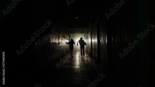 A general shot of a man and a girl running away along a straight corridor from the shadows into the light from a limping man with an ax. High quality 4k footage
