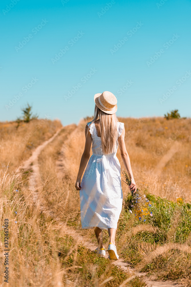 back view of young woman in white dress and straw hat walking in field with bouquet of wildflowers