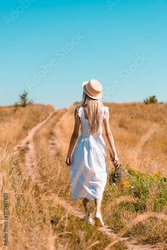 back view of young woman in white dress and straw hat walking in field with bouquet of wildflowers