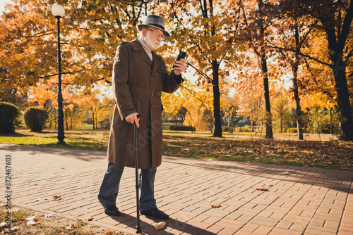 Full length profile photo of retired grey hair grandpa man walk desert park stick read email telephone sunny day use modern technology wear specs long coat headwear autumn colors outdoors