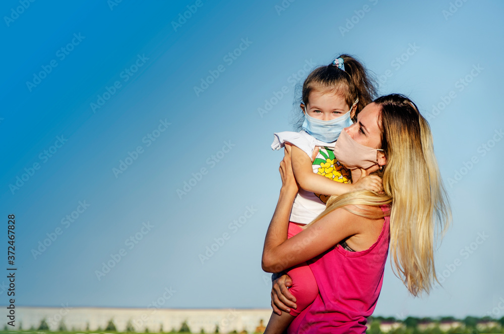 Mom and little daughter in medical masks on a background of blue sky close-up. Face mask on children and adults as a precaution against COVID-19.Portrait photo of mom and daughter against the blue sky Mom and little daughter in medical masks on a background of blue sky close-up. Face mask on children and adults as a precaution against COVID-19.Portrait photo of mom and daughter against the blue sky