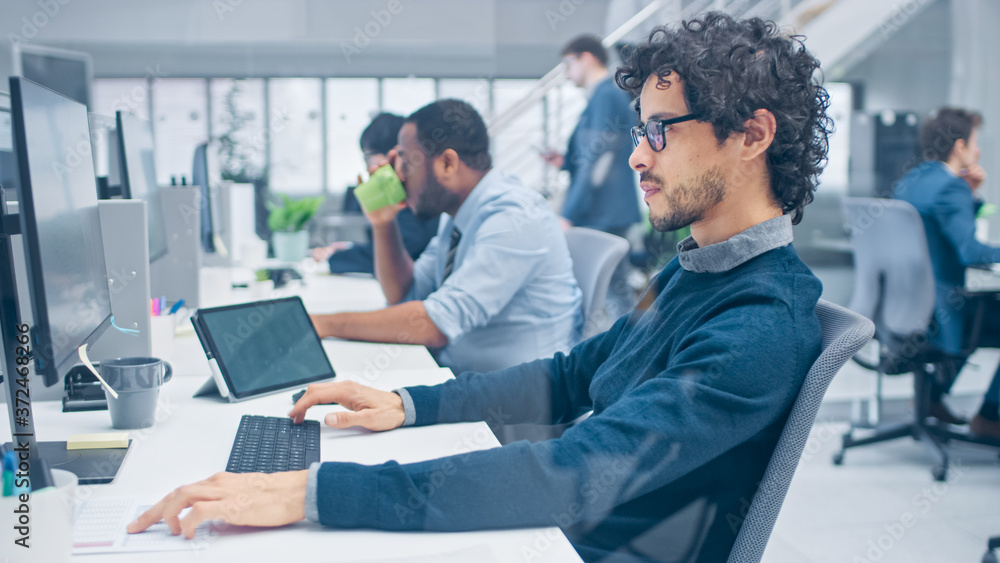Young Handsome Manager with Curly Hair Works on a Desktop Computer and Compares Data with a Software. Diverse and Motivated Business People Work on Computers in Modern Open Office. Young Handsome Manager with Curly Hair Works on a Desktop Computer and Compares Data with a Software. Diverse and Motivated Business People Work on Computers in Modern Open Office.