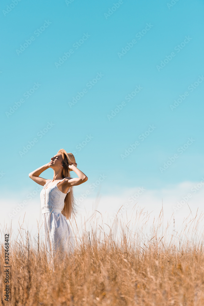 selective focus of stylish woman in white dress touching straw hat while standing with raised head against bule sky selective focus of stylish woman in white dress touching straw hat while standing with raised head against bule sky