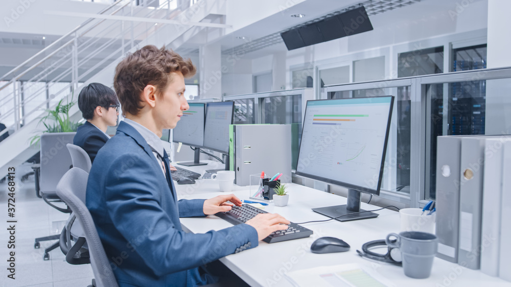 Young Handsome Manager Wearing a Suit and Tie is Working Behind His Desk. Diverse and Motivated Business People Work on Computers in Modern Open Office.