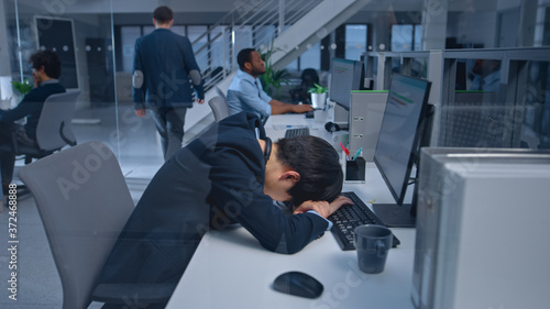 Young Japanese Manager is Tired at his Work Place with Desktop Computer and Falls Asleep. Diverse Multi-ethnic Business People Work on Computers in Modern Open Office.