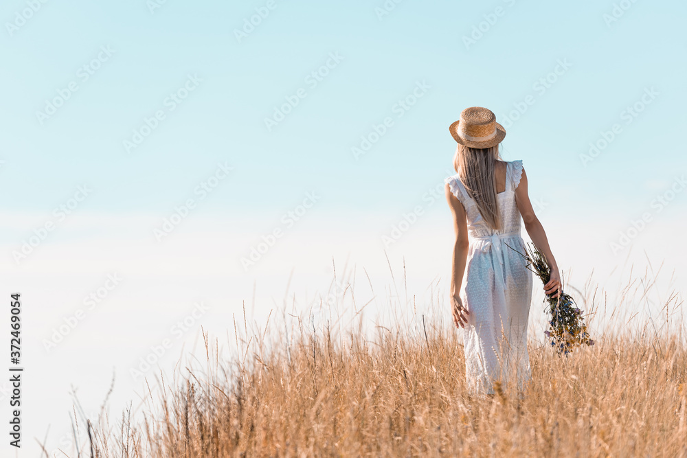 back view of woman in white dress and straw hat holding wildflowers while standing on grassy hill back view of woman in white dress and straw hat holding wildflowers while standing on grassy hill