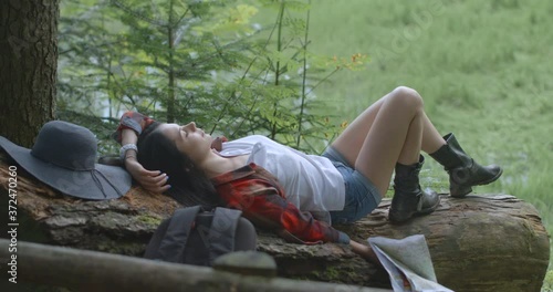 Lying young beautiful hipster with the paper map in hands on the fallen tree. The tourist is tired after hiking in forest. 4k footage. photo