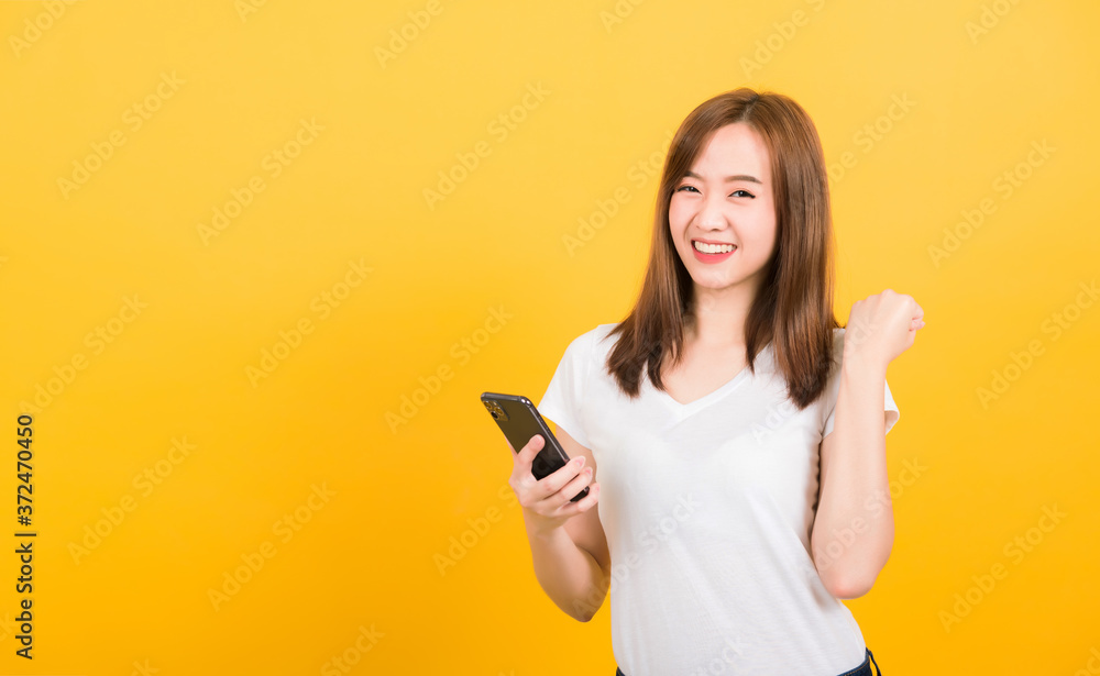 Asian happy portrait beautiful cute young woman teen smiling standing wear t-shirt celebrating win with smart mobile phone looking the phone isolated, studio shot on yellow background with copy space Asian happy portrait beautiful cute young woman teen smiling standing wear t-shirt celebrating win with smart mobile phone looking the phone isolated, studio shot on yellow background with copy space