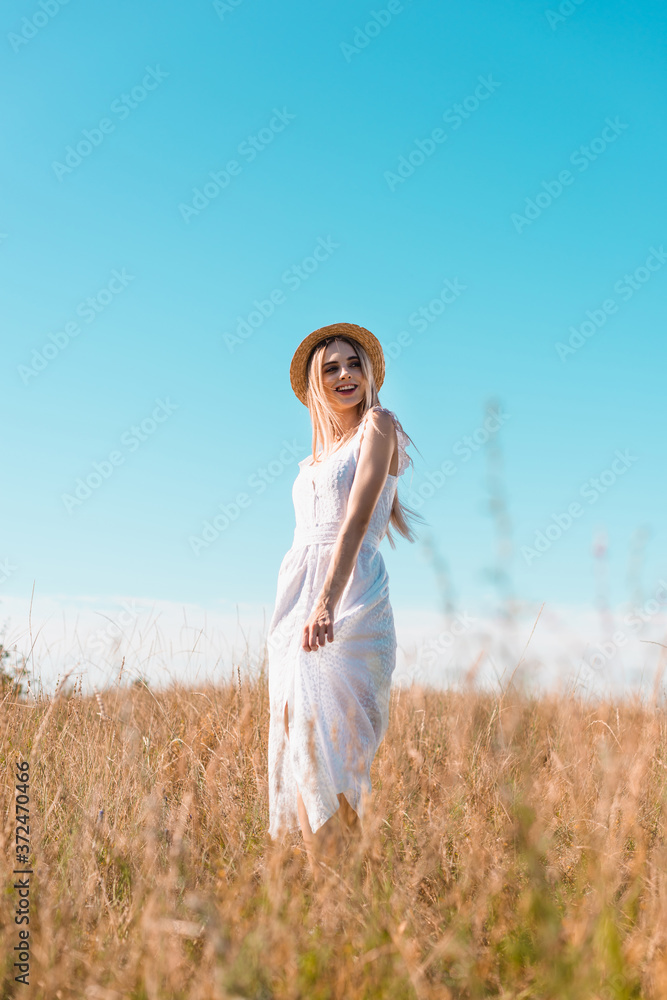 selective focus of blonde woman in white dress and straw hat standing in field against blue sky and looking away selective focus of blonde woman in white dress and straw hat standing in field against blue sky and looking away