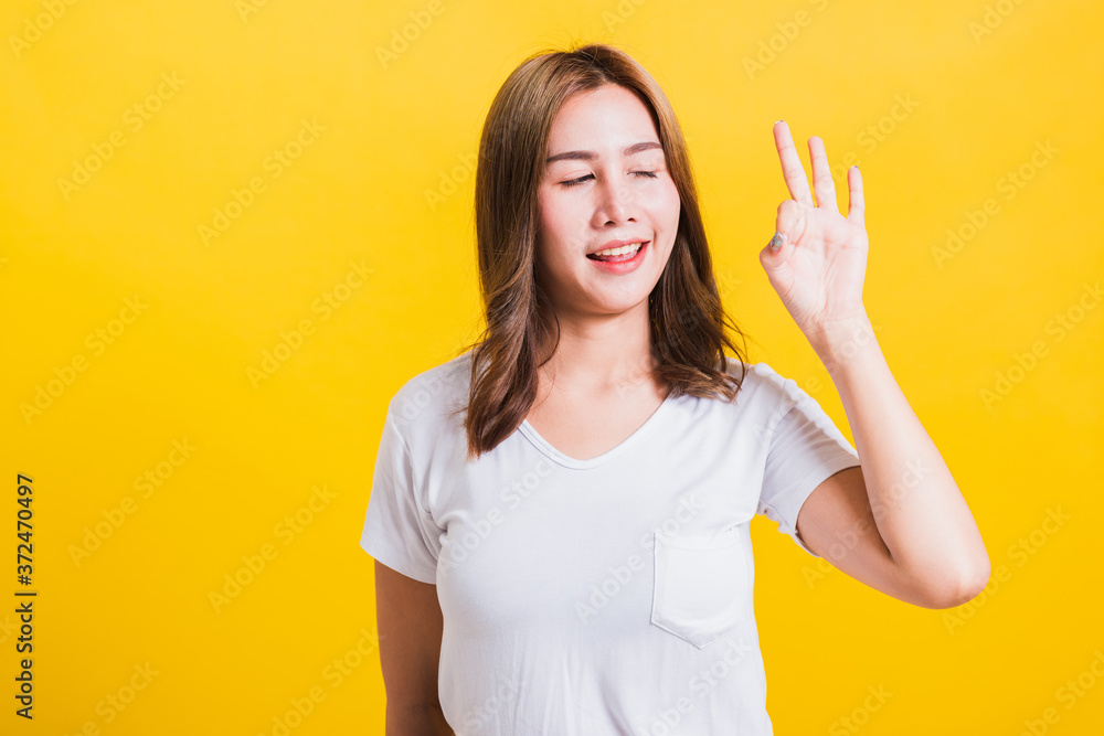 Asian Thai happy portrait beautiful cute young woman standing wear t-shirt showing gesturing ok sign with fingers looking to away, isolated studio shot on yellow background with copy space Asian Thai happy portrait beautiful cute young woman standing wear t-shirt showing gesturing ok sign with fingers looking to away, isolated studio shot on yellow background with copy space