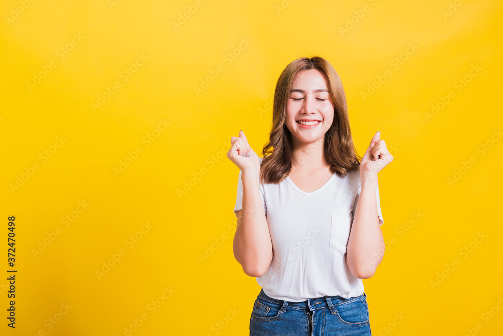 Asian Thai happy portrait beautiful cute young woman standing wear t-shirt makes raised fists up celebrating her winning success she close eyes isolated, studio shot on yellow background Asian Thai happy portrait beautiful cute young woman standing wear t-shirt makes raised fists up celebrating her winning success she close eyes isolated, studio shot on yellow background