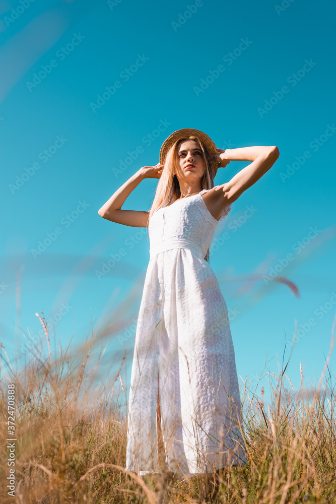 selective focus of young woman in white dress touching straw hat while looking away against blue sky, low angle view