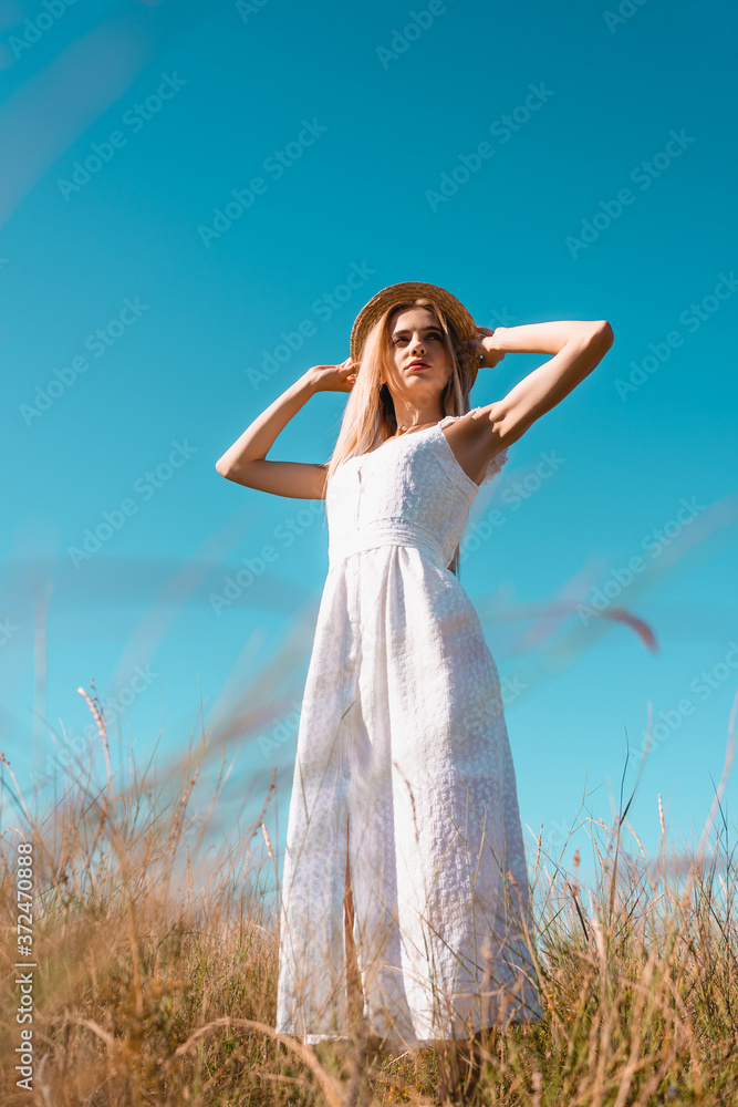 selective focus of young woman in white dress touching straw hat while looking away against blue sky, low angle view selective focus of young woman in white dress touching straw hat while looking away against blue sky, low angle view