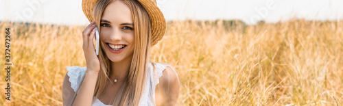panoramic shot of young blonde woman in straw hat looking at camera while talking on smartphone in field