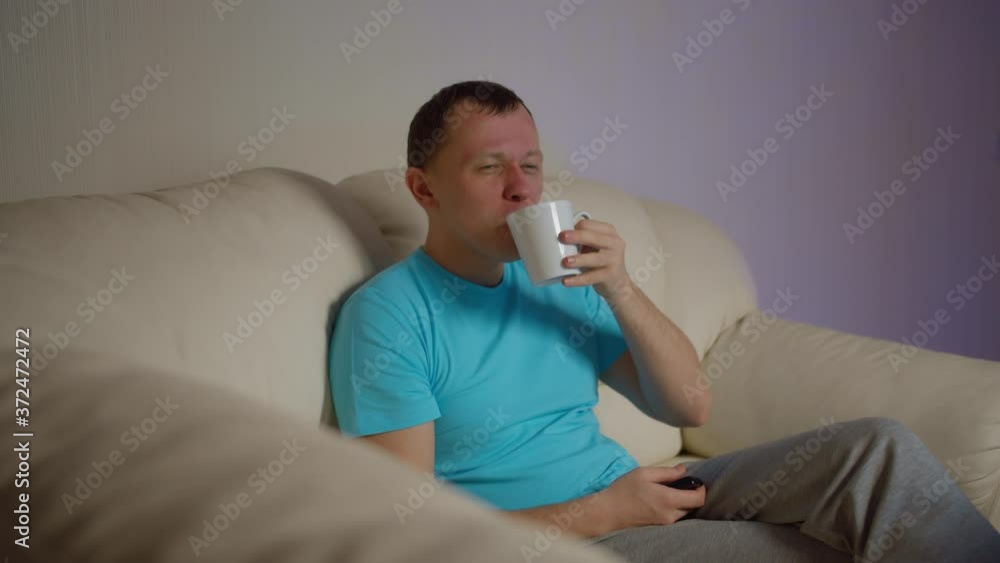 Young man sitting on the sofa in the evening with a mug of tea and watching tv Young man sitting on the sofa in the evening with a mug of tea and watching tv