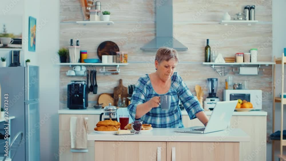 Mature woman typing on laptop in kitchen during breakfast and driking coffee. Elderly retired person working from home, telecommuting using remote internet job online communication on modern Mature woman typing on laptop in kitchen during breakfast and driking coffee. Elderly retired person working from home, telecommuting using remote internet job online communication on modern