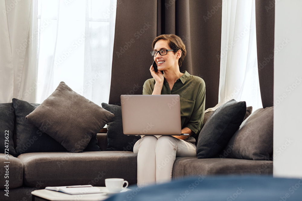 Smiling busineswoman sitting on sofa and talking on phone in hotel room. Young entrepreneur working from apartment. Smiling busineswoman sitting on sofa and talking on phone in hotel room. Young entrepreneur working from apartment.