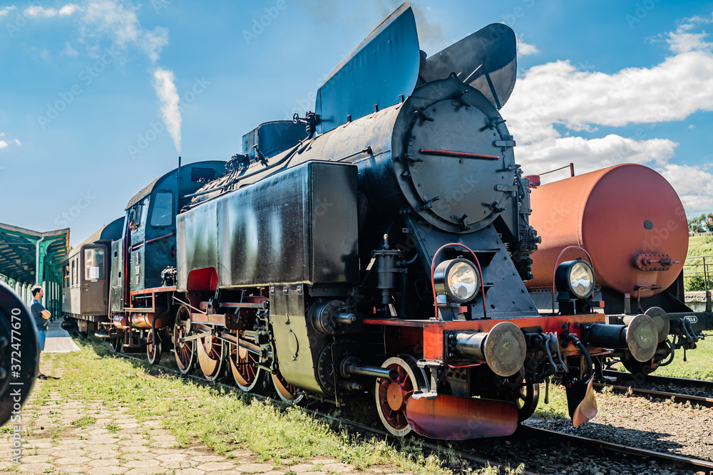 Obraz premium Huge vintage steam locomotive, red painted steel wheel detail close up. Coal-powered steam train stands on a siding. Classic gigantic heavy railway machinery. Side view of power parts of machine.