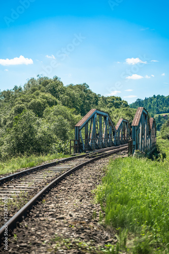 Railway bridge over river in Polish mountain. Heavy rusty steel old industrial train overpass in natural scenic landscape, blue summer sky.