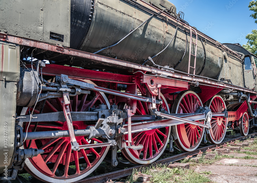 Naklejka premium Huge vintage steam locomotive, red painted steel wheel detail close up. Coal-powered steam train stands on a siding. Classic gigantic heavy railway machinery. Side view of power parts of machine.