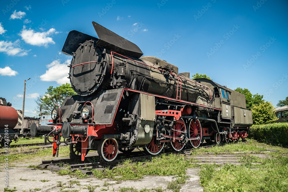 Naklejka premium Huge vintage steam locomotive, red painted steel wheel detail close up. Coal-powered steam train stands on a siding. Classic gigantic heavy railway machinery. Side view of power parts of machine.