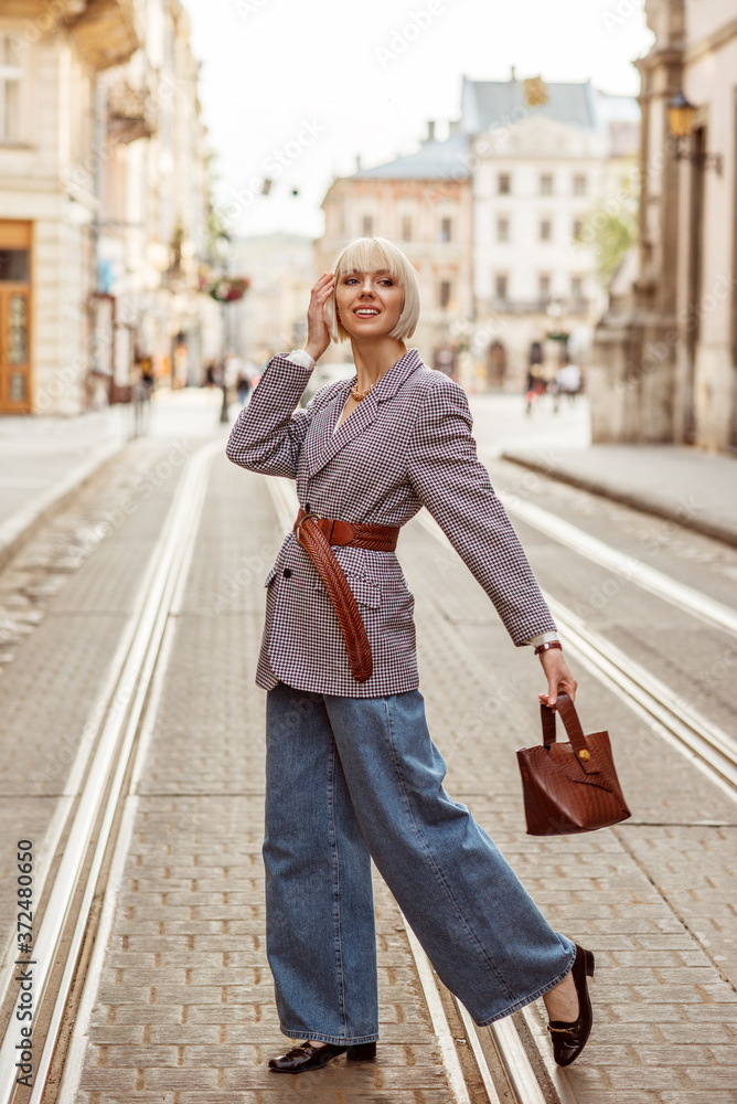 Street autumn fashion portrait of elegant happy smiling woman wearing trendy checkered blazer, wide belt, wide leg jeans, holding brown top handle bag, walking in street of European city
Street autumn fashion portrait of elegant happy smiling woman wearing trendy checkered blazer, wide belt, wide leg jeans, holding brown top handle bag, walking in street of European city