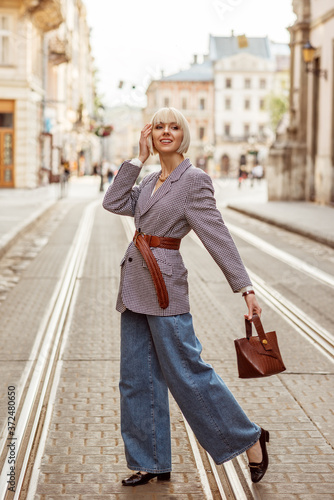 Street autumn fashion portrait of elegant happy smiling woman wearing trendy checkered blazer, wide belt, wide leg jeans, holding brown top handle bag, walking in street of European city 