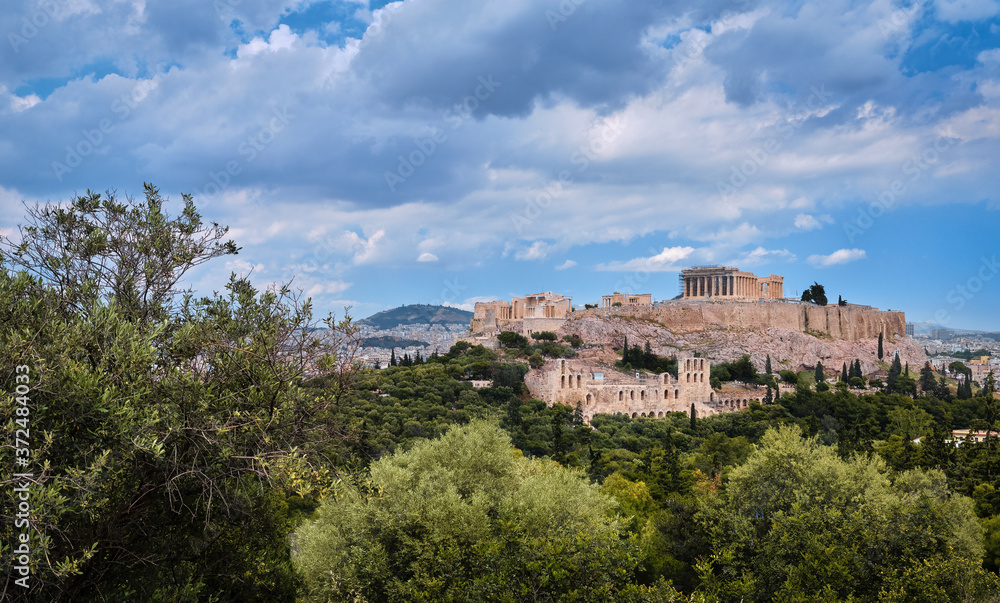 View of Acropolis hill and theater of Odeon in Athens, Greece from the hill of Philoppapos or Muses in summer daylight with great clouds in blue sky.