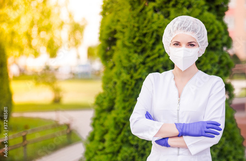 A beautiful girl in a medical suit, a protective mask on her face, gloves and a medical cap poses against a background of greenery. The girl stands with crossed arms and looks into the camera.